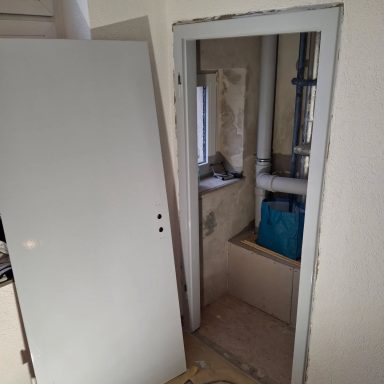 Ein offener Raum mit einer Tür, einem Fenster und sichtbaren Rohren in der Ecke.