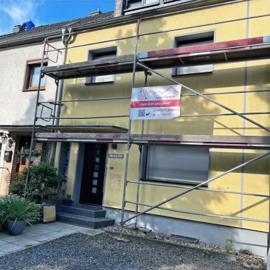 Gelbes Haus mit Gerüst und Renovierungsschild, Pflanzen vor dem Eingang.