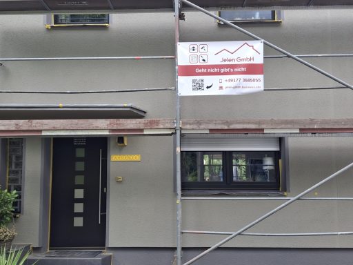Fassade eines Hauses mit Gerüst und Werbebanner, daneben ein Eingang mit schwarzer Tür.